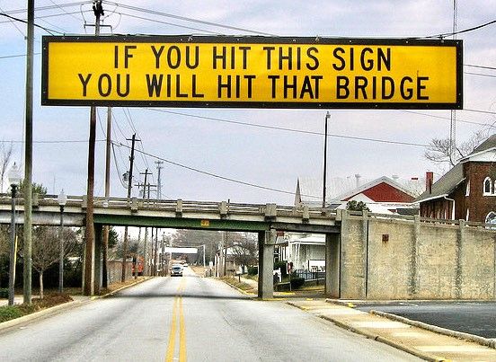Placa amarela com letras pretas, em frente a uma ponte, com a frase "If you hit this sign, you will hit that bridge".Em Português: "Se você bater nessa placa, vai bater naquela ponte."