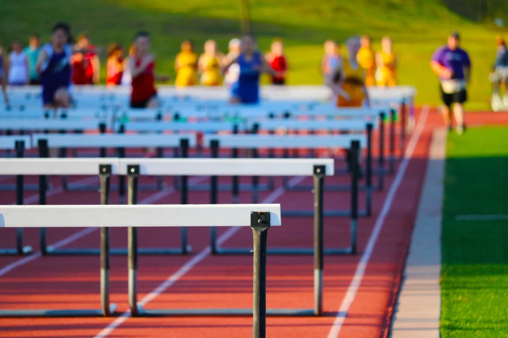 Uma pista de atletismo com as barreiras perfiladas, e alguns atletas iniciando uma competição ao fundo, com o segundo plano desfocado.