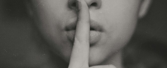 Foto em preto e branco, mostrando parte do rosto de uma mulher, com o dedo indicador na vertical sobre os lábios, fazendo o gesto de "Silêncio".