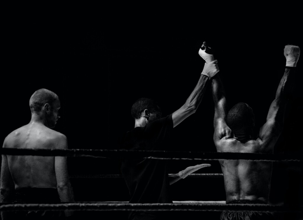Foto em preto e branco mostrando dois lutadores de costas, em um ringue de boxe, após uma luta, com o juiz levantando o braço do vencedor, à direita, enquanto o perdedor observa.
