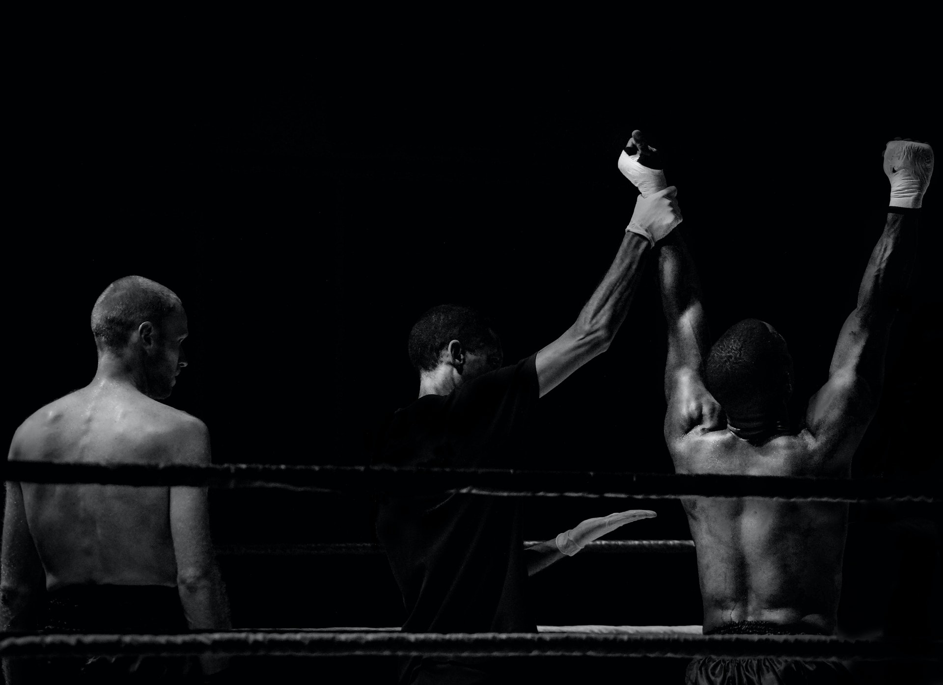 Foto em preto e branco mostrando dois lutadores de costas, em um ringue de boxe, após uma luta, com o juiz levantando o braço do vencedor, à direita, enquanto o perdedor observa. É uma analogia às pessoas que, ao negociar, preferem competir em vez de cooperar - o que sempre gera um perdedor.