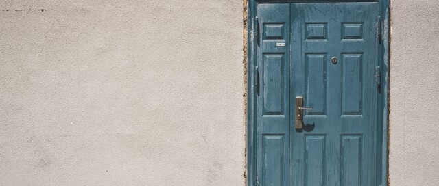 Uma porta de madeira azul celeste, com uma pintura em efeito de pátina, na frente de uma parede bege, de cimento.