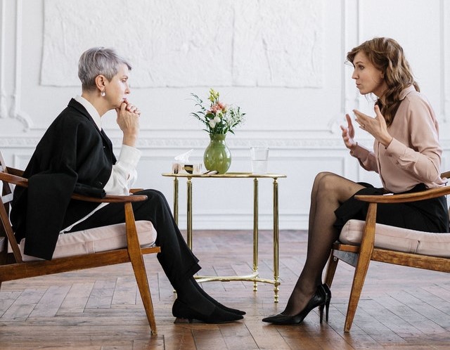 Duas mulheres sentadas em poltronas de madeira, frente à frente, conversando assuntos profissionais.