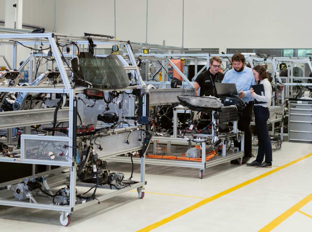 Interior de uma fábrica, com vários equipamentos distribuídos sobre um chão claro e demarcado com fitas amarelas. No lado direito da foto, dois homens e uma mulher analisam informações de um notebook.