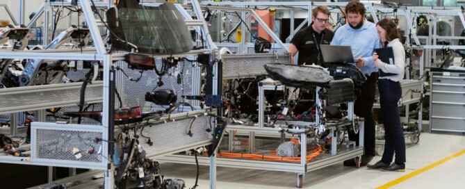 Interior de uma fábrica, com vários equipamentos distribuídos sobre um chão claro e demarcado com fitas amarelas. No lado direito da foto, dois homens e uma mulher analisam informações de um notebook.