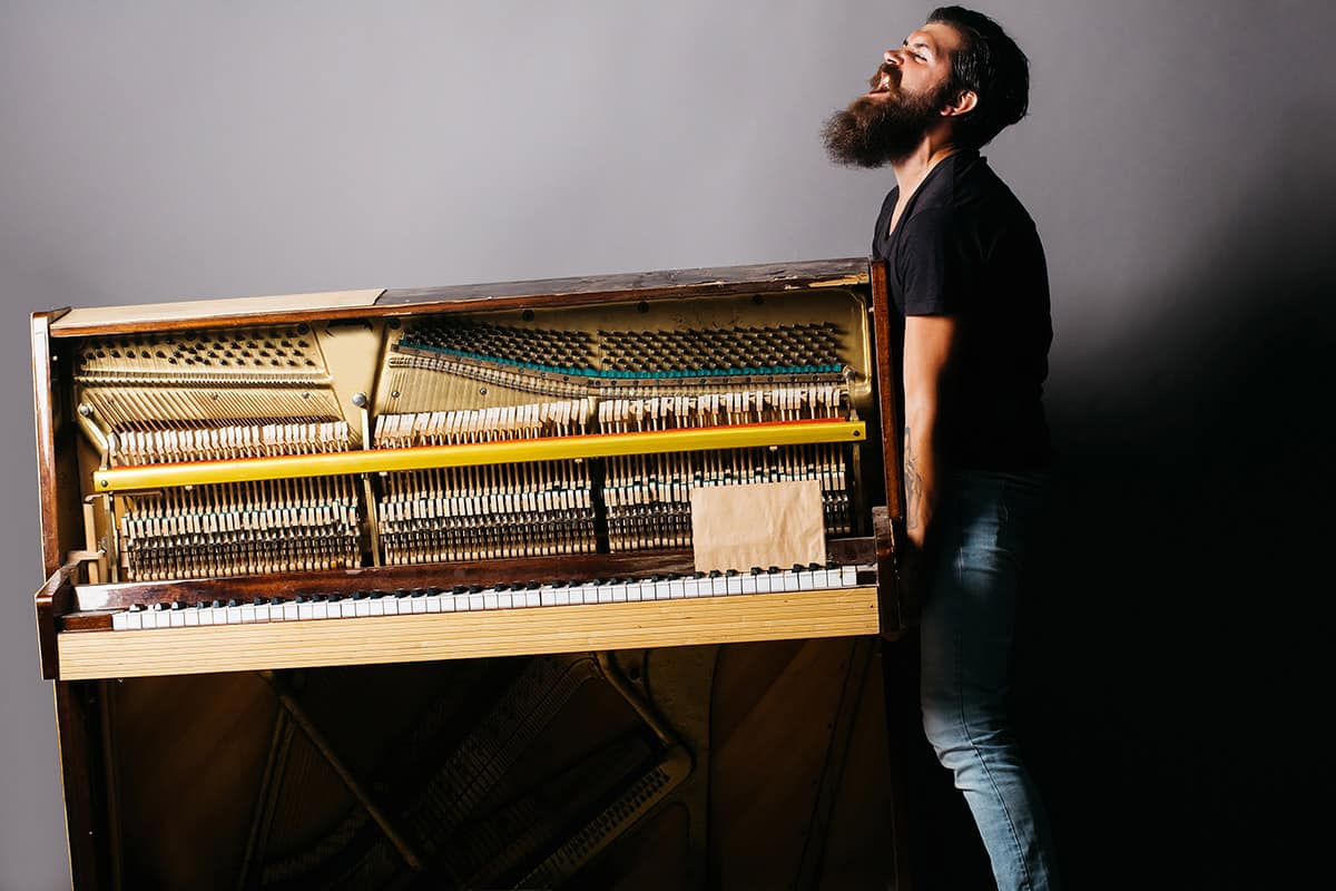 Homem com barba comprida, camiseta preta e calça jeans aparece carregando um piano sozinho, com uma expressão de quem está fazendo muito esforço.