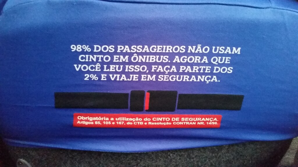 Foto da parte de trás do encosto de cabeça do banco de um ônibus intermunicipal, com um aviso onde se lê: “98% dos passageiros não usam cinto em ônibus. Agora que você leu isso, faça parte dos 2% e viaje em segurança.”