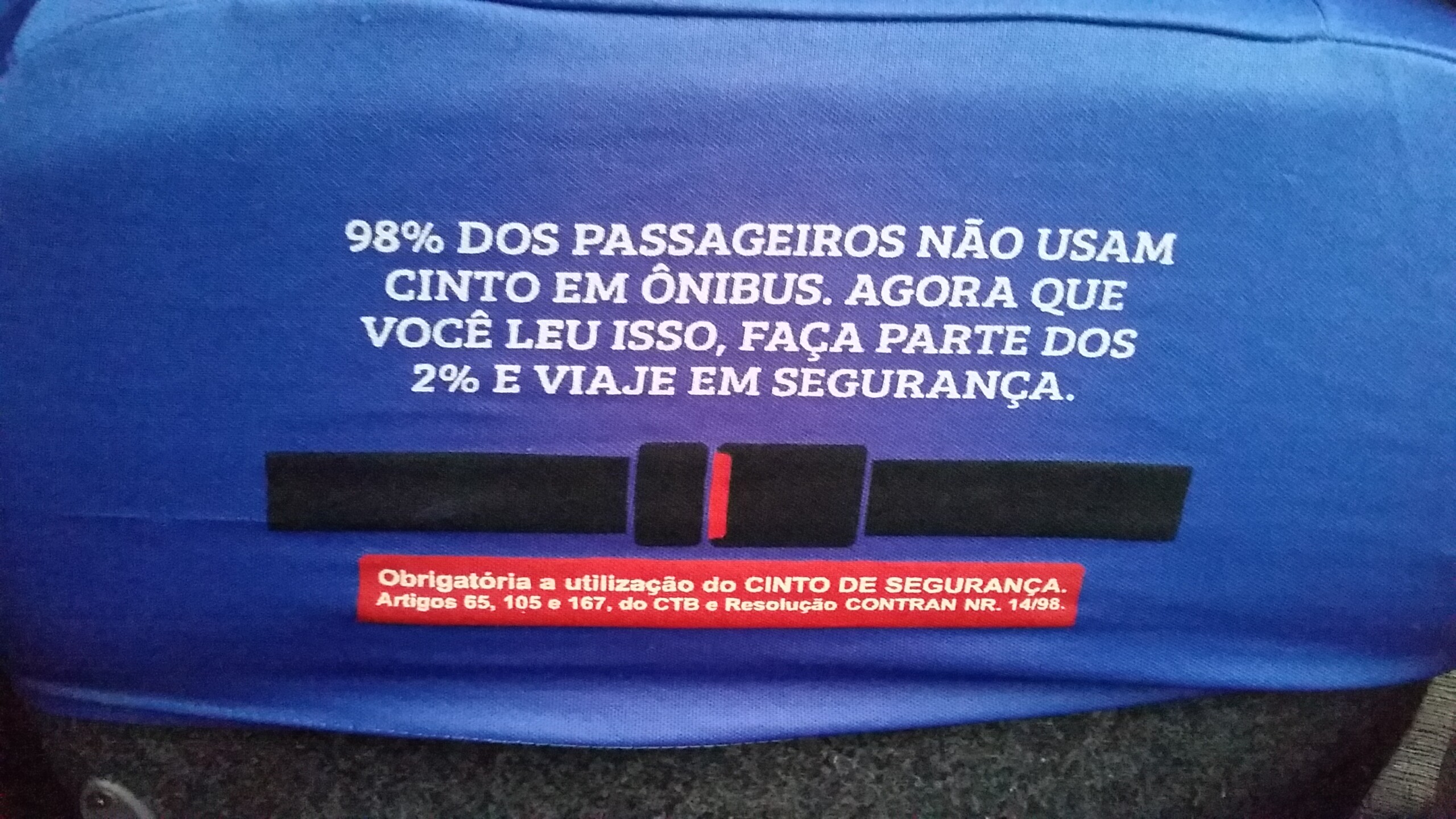 Foto da parte de trás do encosto de cabeça do banco de um ônibus intermunicipal, com um aviso onde se lê: “98% dos passageiros não usam cinto em ônibus. Agora que você leu isso, faça parte dos 2% e viaje em segurança.”