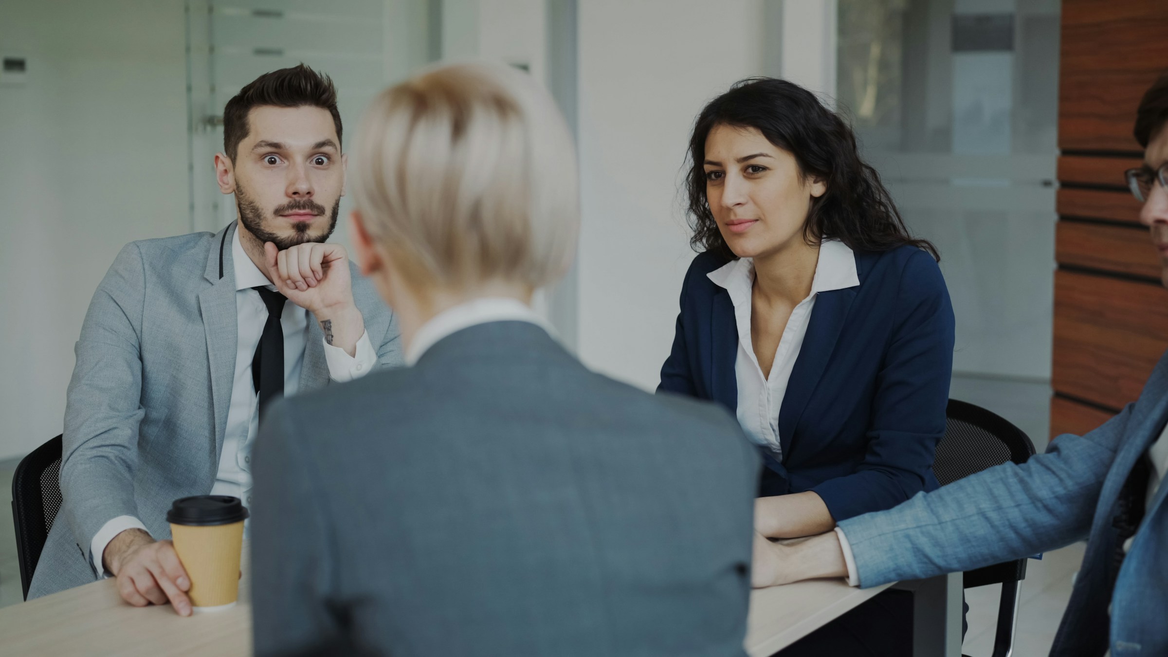 Quatro pessoas em uma sala de reunião de uma empresa. De costas, uma mulher loira de cabelos curtos diz algo que surpreende as outras pessoas. De frente para ela, um rapaz de terno cinza, camisa branca e gravata preta faz uma expressão de espando, com os olhos arregalados. Ao seu lado, uma mulher jovem olha fixamente com uma expressão de dúvida. A quarta pessoa é um homem de meia idade, cabelos curtos e óculos, mas só aparece metade do seu corpo.