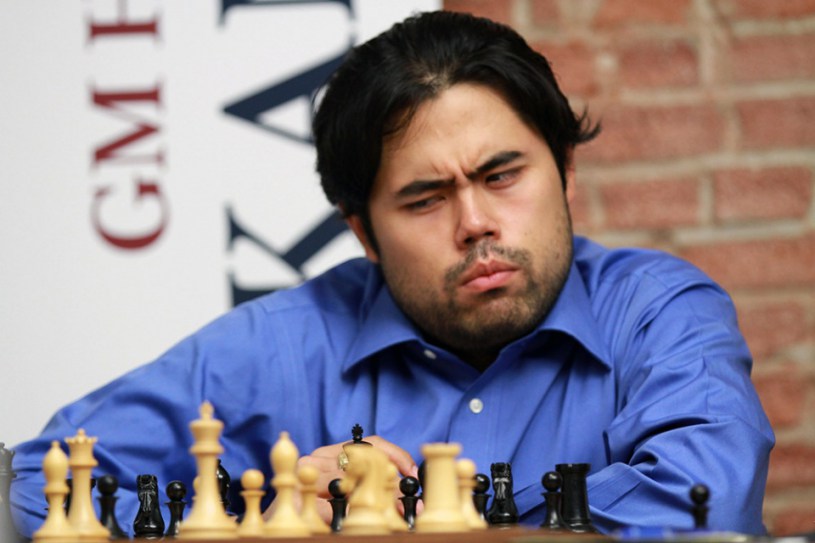 O Grande Mestre americano de xadrez Hikaru Nakamura analisa uma posição diante de um tabuleiro com peças de xadrez. Ele é um homem asiático de cabelos escuros, curtos, repartidos no meio e usa uma camisa azul celeste. Sua expressão facial demonstra desconfiança.