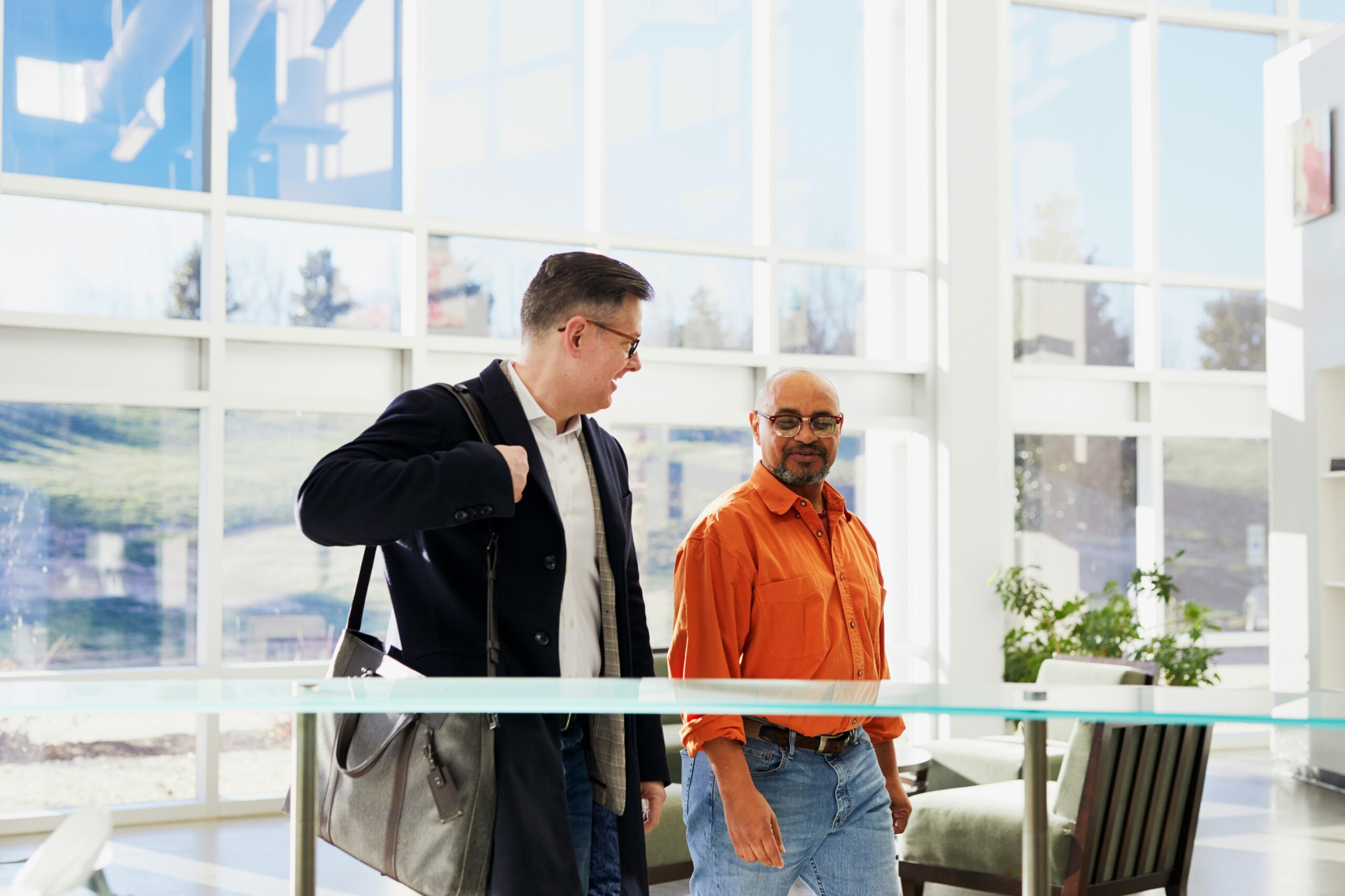 Duas pessoas caminhando pelo lobby de um edifício. À esquerda um homem branco, alto, de cabelos escuros, curtos e óculos, vestindo calça azul, camisa branca e um sobretudo azul escuro, carregando uma pasta de alça, à tiracolo. À direita um homem mulato, careca e com cavanhaque grisalho, também de óculos, vestindo uma calça jeans azul claro e uma camisa laranja abotoada e com as mangas arregaçadas.