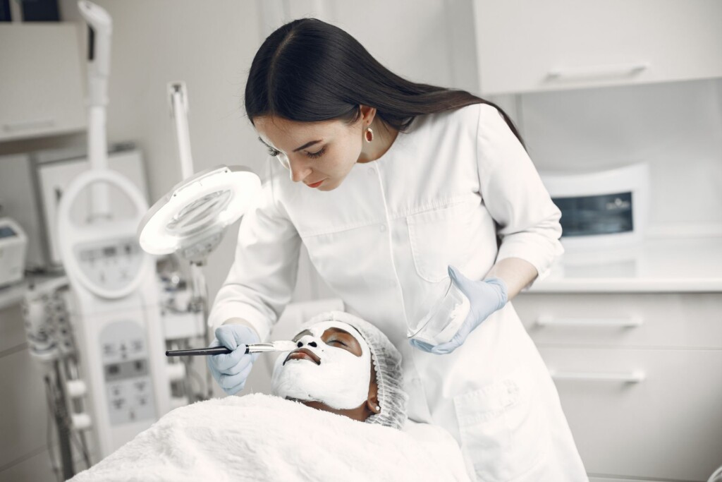 Uma mulher de pé, vestindo um jaleco branco cobre com uma máscara de creme o rosto de uma paciente, que está deitada sobre uma maca, com um tecido branco sobre o corpo. As duas estão no interior de uma clínica de estética com paredes e móveis todos brancos, além de vários equipamentos médicos.