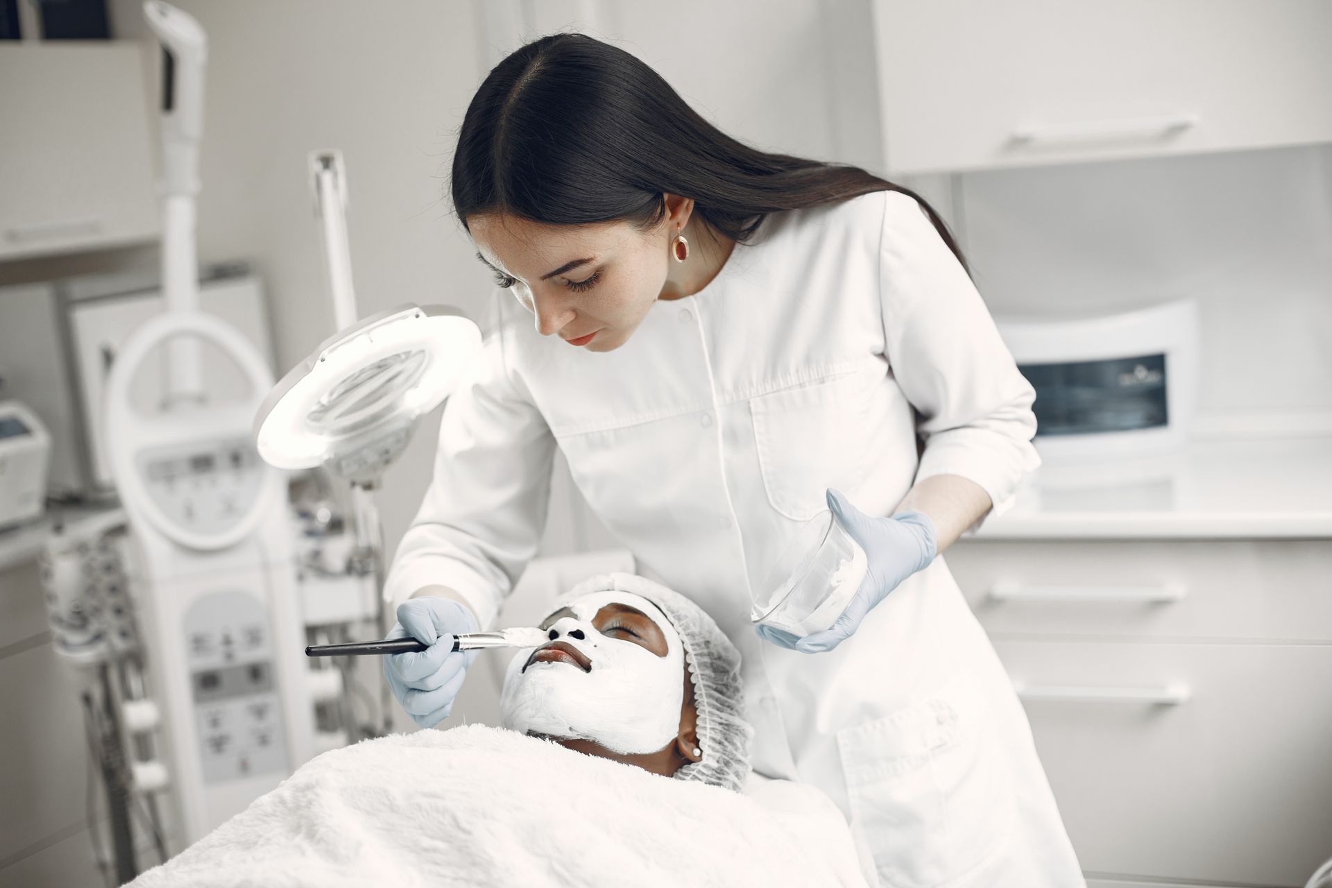 Uma mulher de pé, vestindo um jaleco branco cobre com uma máscara de creme o rosto de uma paciente, que está deitada sobre uma maca, com um tecido branco sobre o corpo. As duas estão no interior de uma clínica de estética com paredes e móveis todos brancos, além de vários equipamentos médicos.