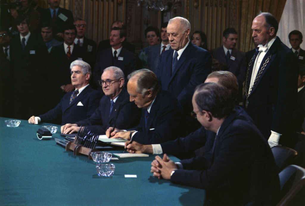 A assinatura do Acordo de Paz de Paris, pelo secretário de estado americano William Rogers, em 27/01/1973. Na sala estão várias pessoas de terno e gravata de pé, enquanto que cinco diplomatas sentam-se em volta de uma mesa redonda, com o documento sendo assinado pelo que está no meio.